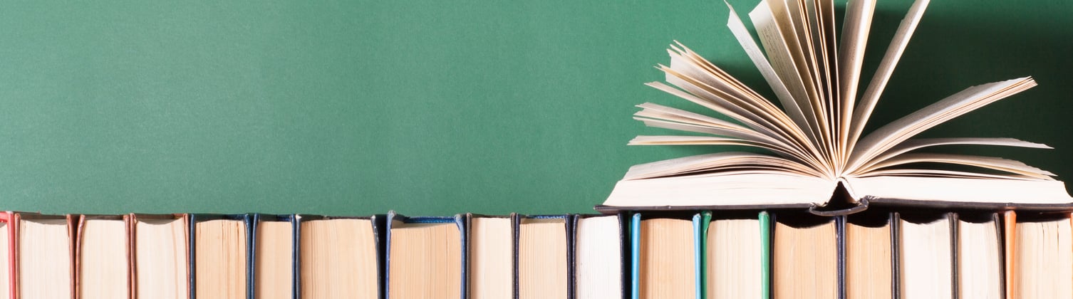 A row of books with the open edge facing front, and one singular book placed on top of them with open pages, fanned out.