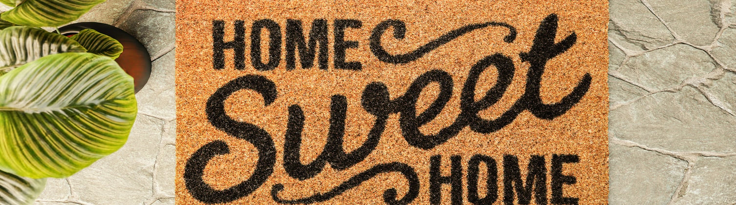 A doormat outside someones front door with a plant in the corner. The mat says the words 