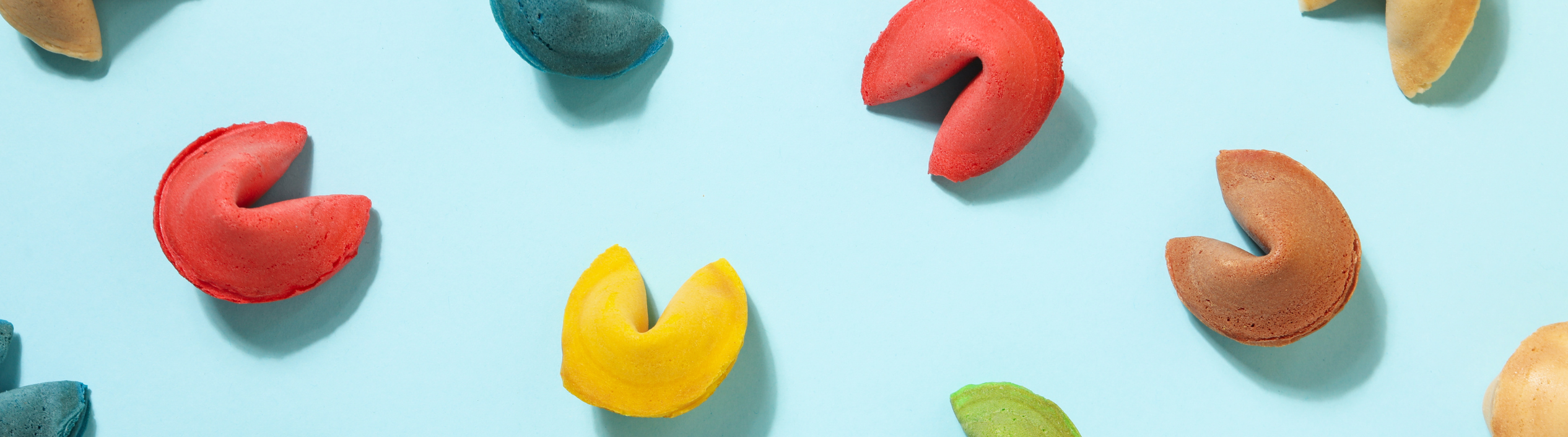 Multiple yellow, green, brown , red and teal fortune cookies arranged in a neat grid on a light blue surface.