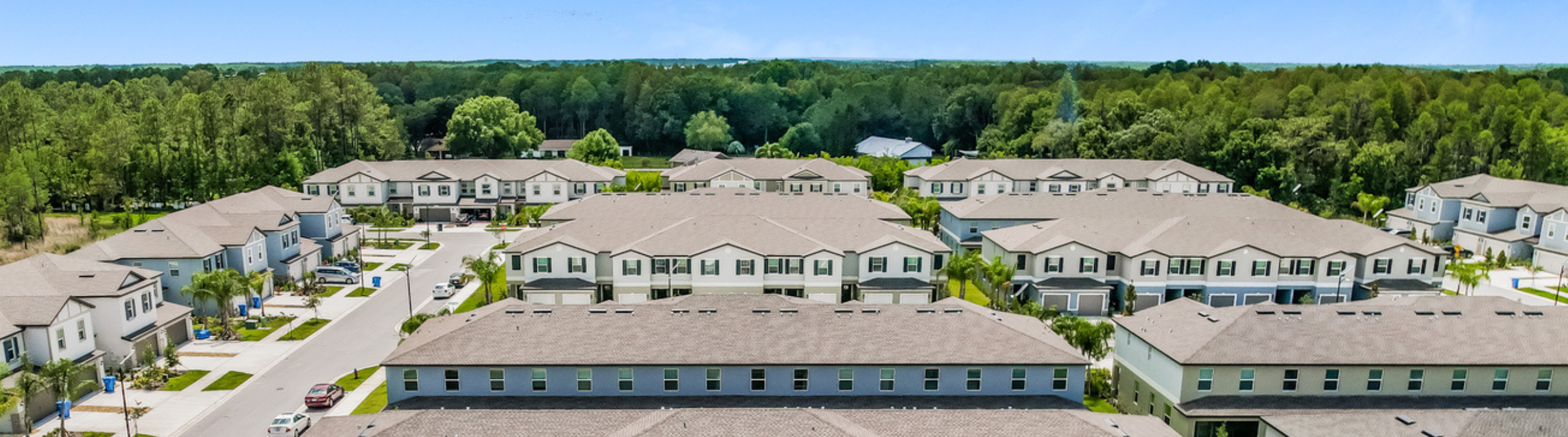 An arial shot of a community of multiple row houses with a lot of trees and blue sky in the background.