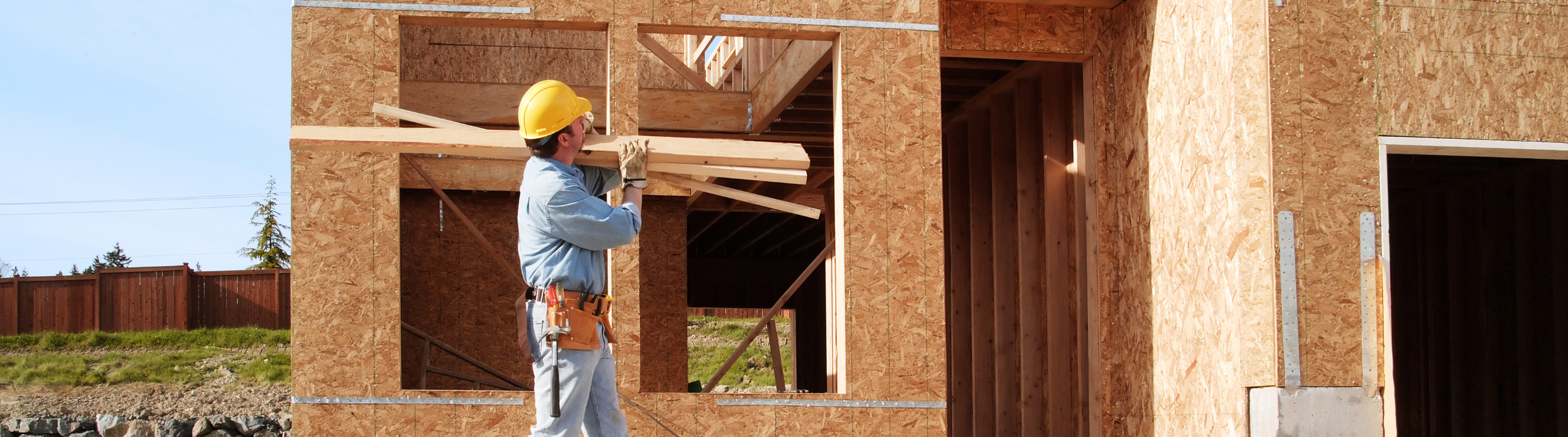A man in a yellow hard hat carrying multiple 2 x 4s in a house construction setting.