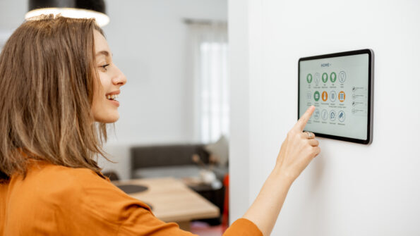Young woman controlling home with a digital touch screen panel installed on the wall in the living room.